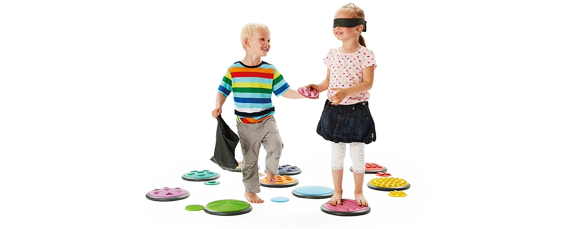 Zwei Kinder gehen barfu&szlig; &uuml;ber bunte Tastscheiben mit unterschiedlichen Oberfl&auml;chen, das M&auml;dchen tr&auml;gt eine Augenbinde, der Junge reicht ihm eine Scheibe